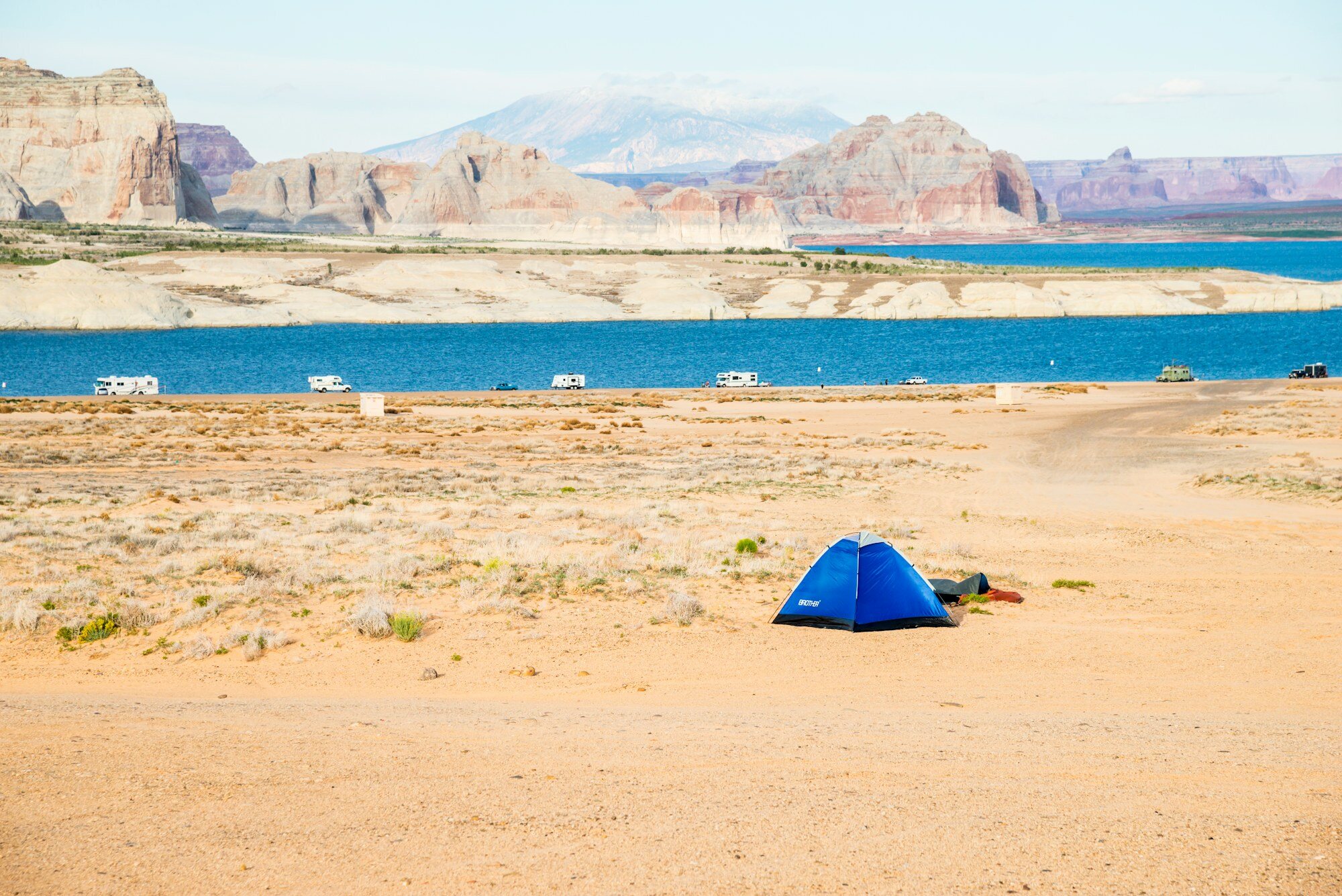 Desert camping tent set up on sandy shore beside blue reservoir with red rock formations in background